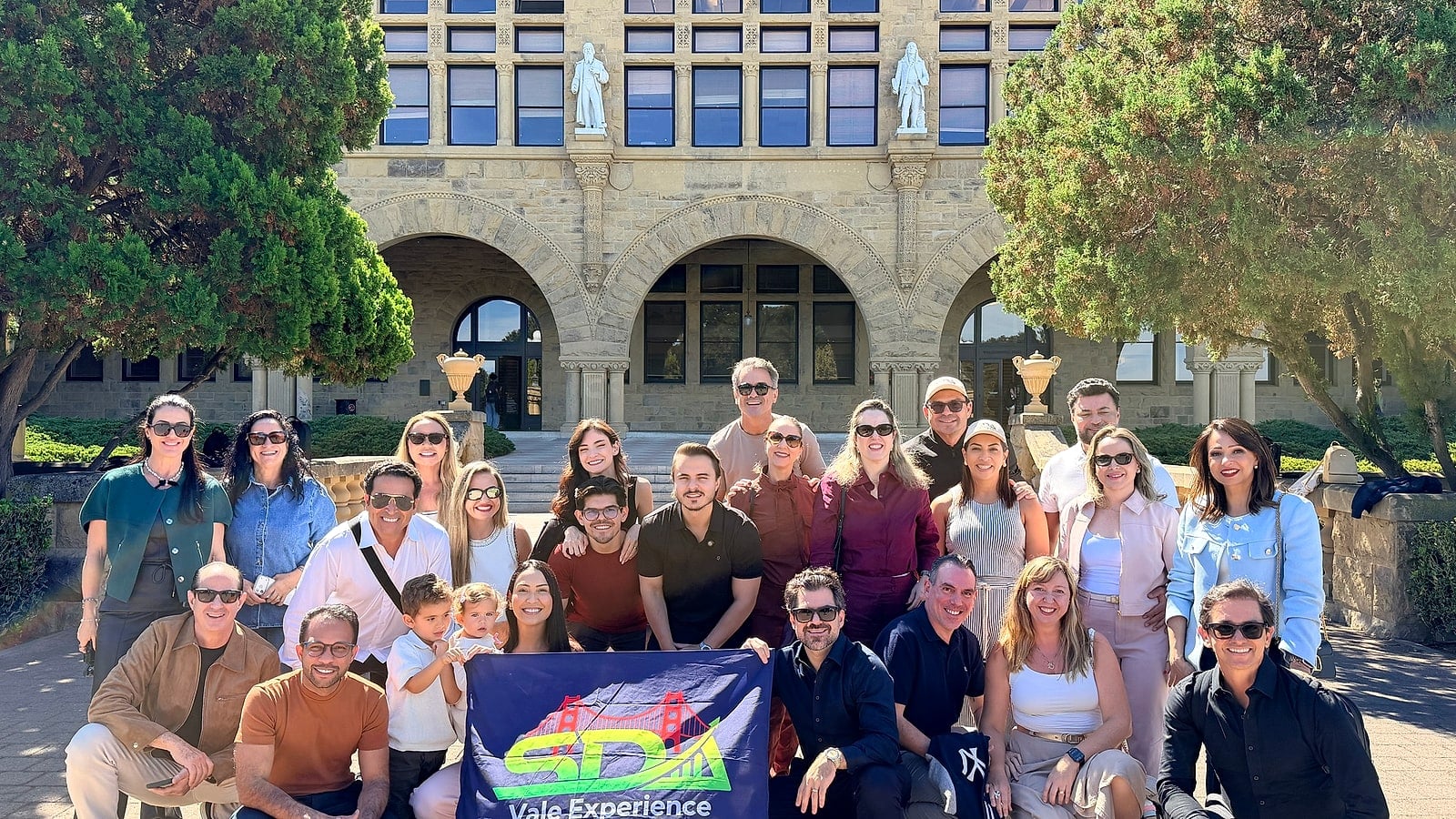 Grupo do SDA Vale Experience reunido diante de um dos prédios históricos do campus de Stanford, em Palo Alto, com a bandeira do grupo