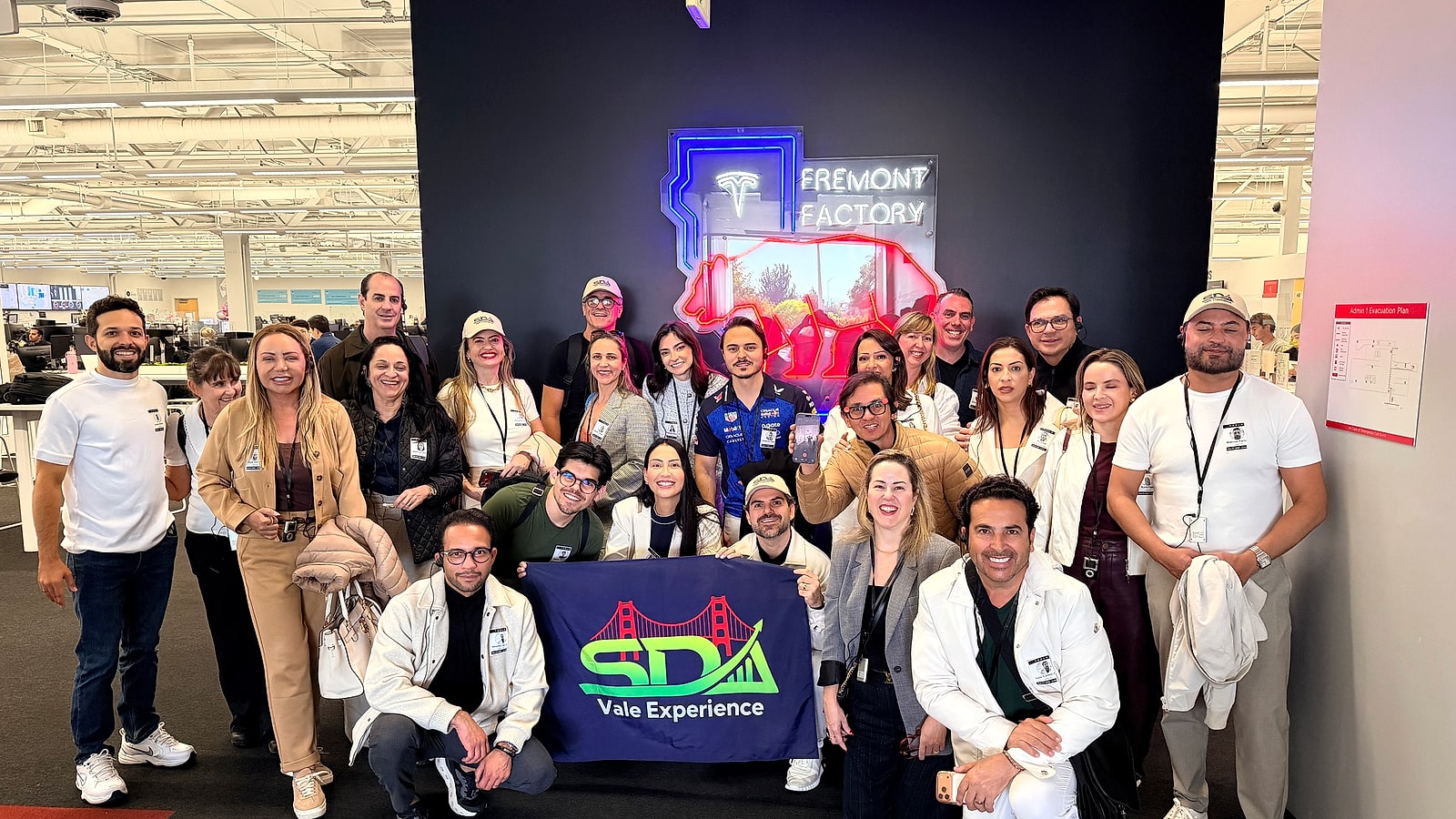 Grupo do SDA Vale Experience reunido diante do letreiro em neon da Tesla Fremont Factory, segurando a bandeira do grupo
