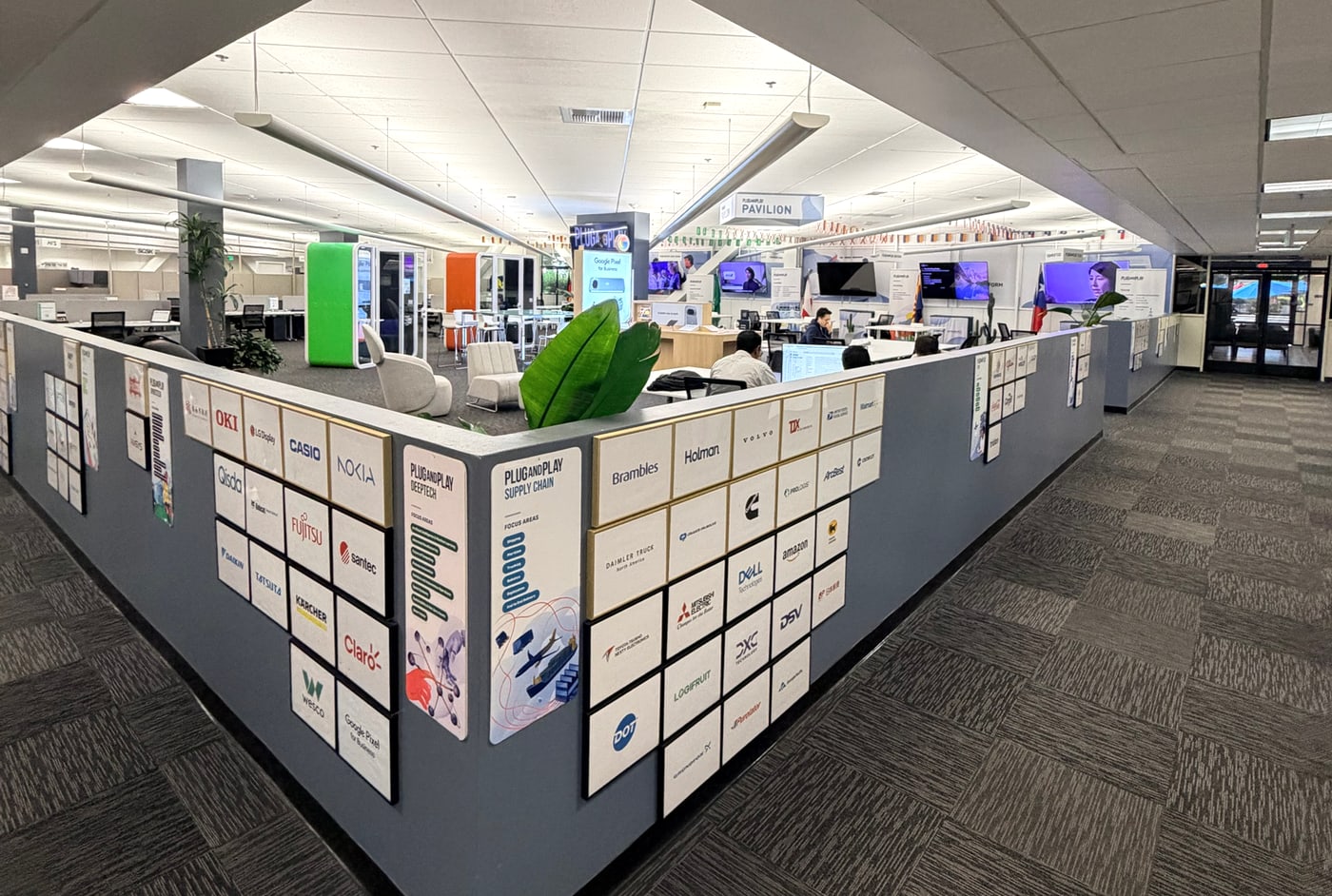 Parede no escritório do Plug and Play coberta de logotipos de corporações parceiras, entre elas Nokia, Casio, Fujitsu, Volvo, Amazon, Dell, Daimler Truck, Mitsubishi Electric, Cummins, Prologis e Claro