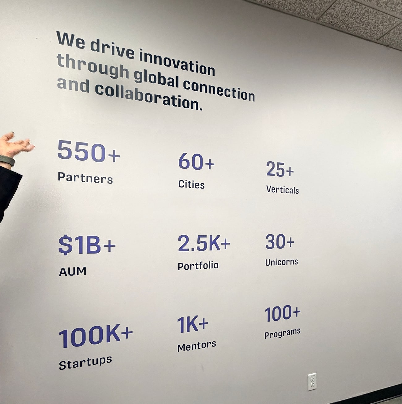 Parede do Plug and Play com os números da empresa: 550+ parceiros, 60+ cidades, 25+ verticais, US$ 1 bilhão+ sob gestão, 2,5 mil+ de portfólio, 30+ unicórnios, 100 mil+ startups, 1 mil+ mentores e 100+ programas