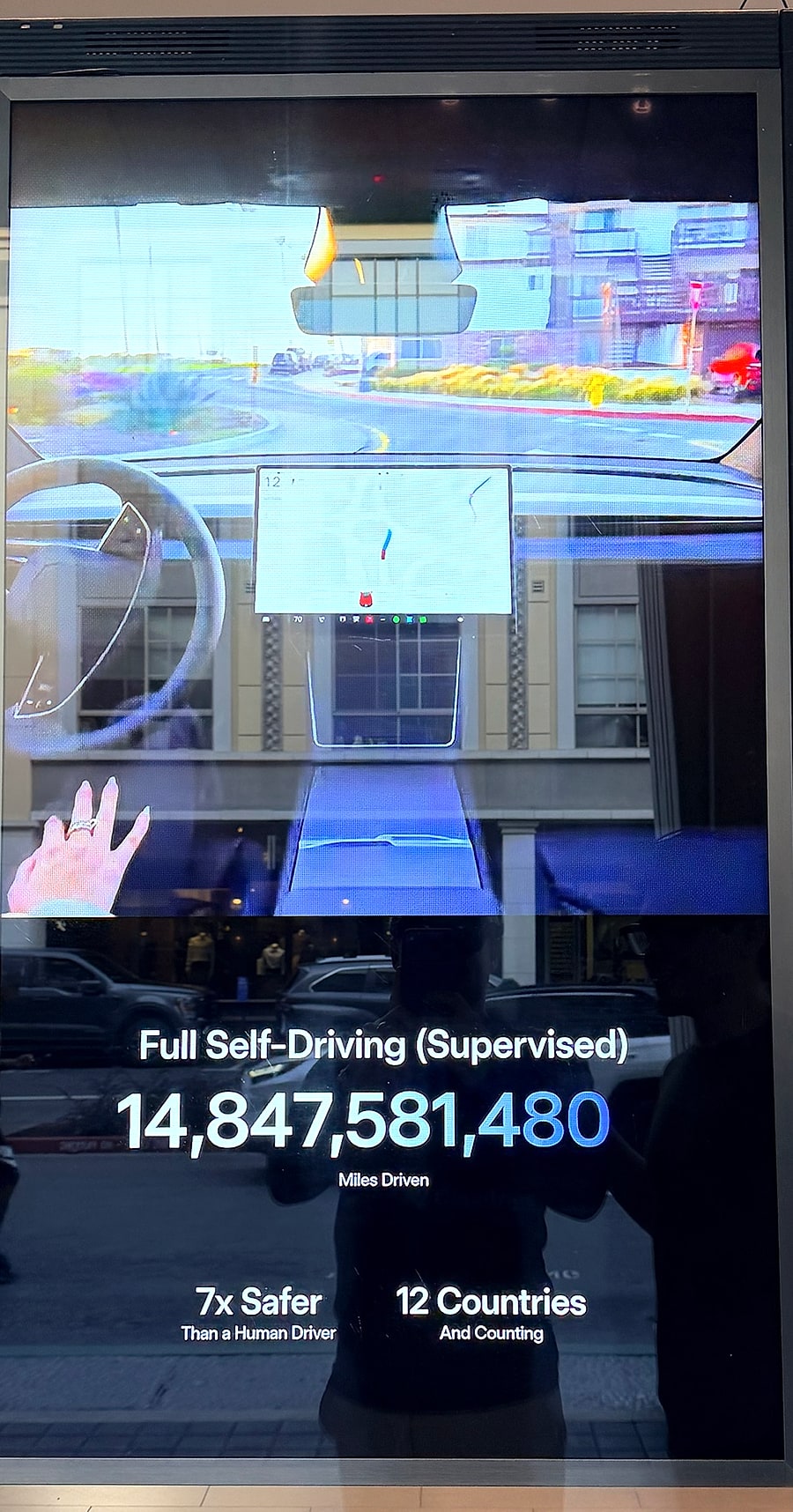 Painel em loja da Tesla mostrando o contador do Full Self-Driving Supervised com 14.847.581.480 milhas dirigidas, além de 7x mais seguro que um motorista humano e 12 países