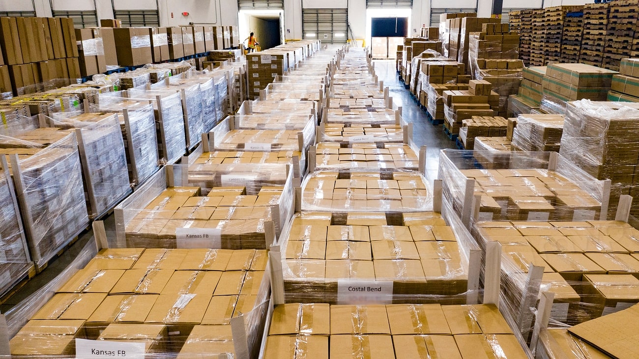 Corredor de um centro de distribuição com fileiras de paletes de caixas de papelão embaladas, vistas em perspectiva