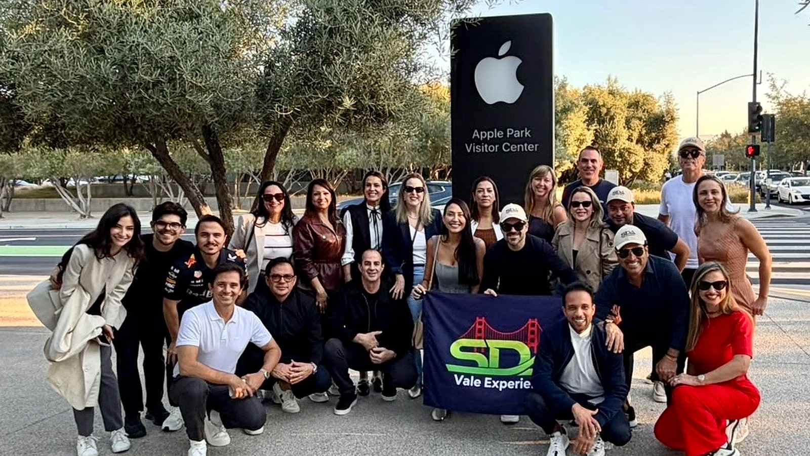 Grupo do SDA Vale Experience reunido diante da placa do Apple Park Visitor Center, em Cupertino, segurando a bandeira do grupo