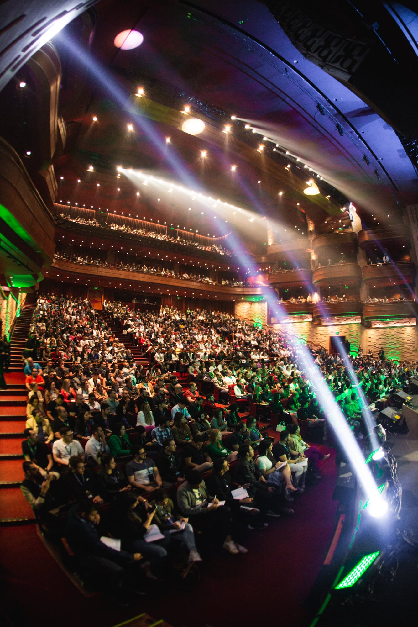 Vista em grande angular do teatro lotado, com feixes de luz verde e branca sobre a plateia